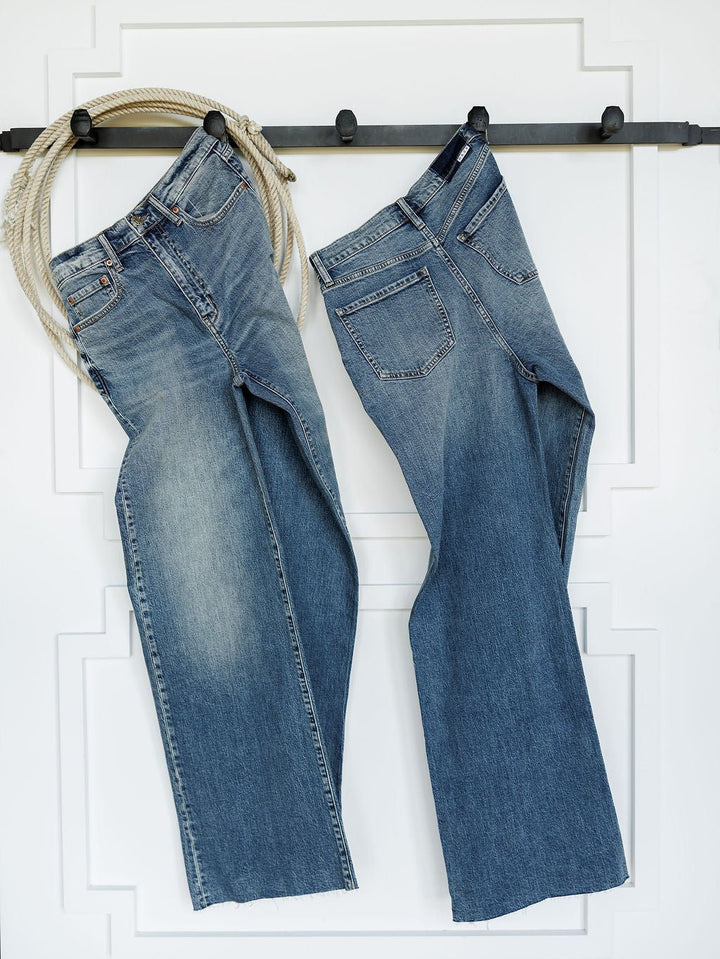 Two pairs of The Leyden High Rise Wide Leg Jean by Savannah Sevens western life{&}style hang on a black wall hook, with a coiled rope draped over the left hook. A white wall with decorative molding serves as the background.