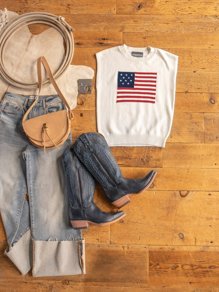 Flat lay of The Liberty American flag top by Savannah Sevens western life{&}style, light blue jeans, blue cowboy boots, a tan crossbody bag, coiled rope, and cowhide rug arranged on a wooden floor.