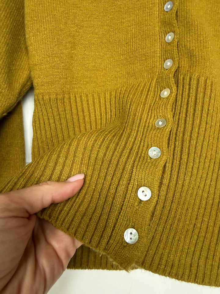 A hand holds the ribbed hem of the Lindsborg Chartreuse Button Up Cardigan by Savannah Sevens western life{&}style, displaying a row of shiny silver buttons along the edge.