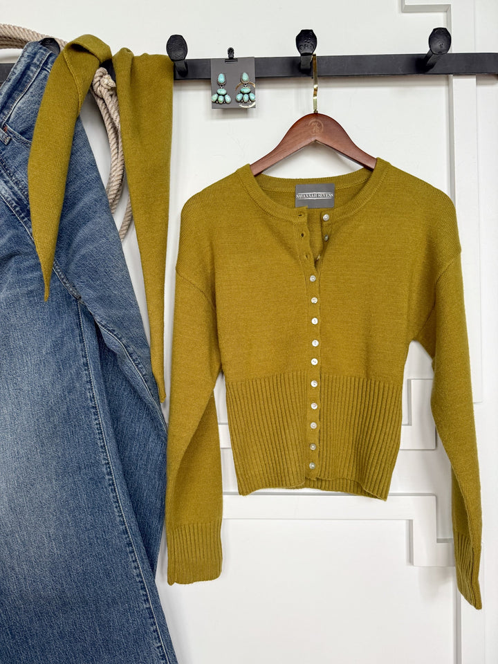The Lindsborg Chartreuse Button Up Cardigan by Savannah Sevens western life{&}style hangs on a wooden hanger beside denim jeans, a matching belt, a rope, and turquoise earrings displayed on black hooks against a white wall.