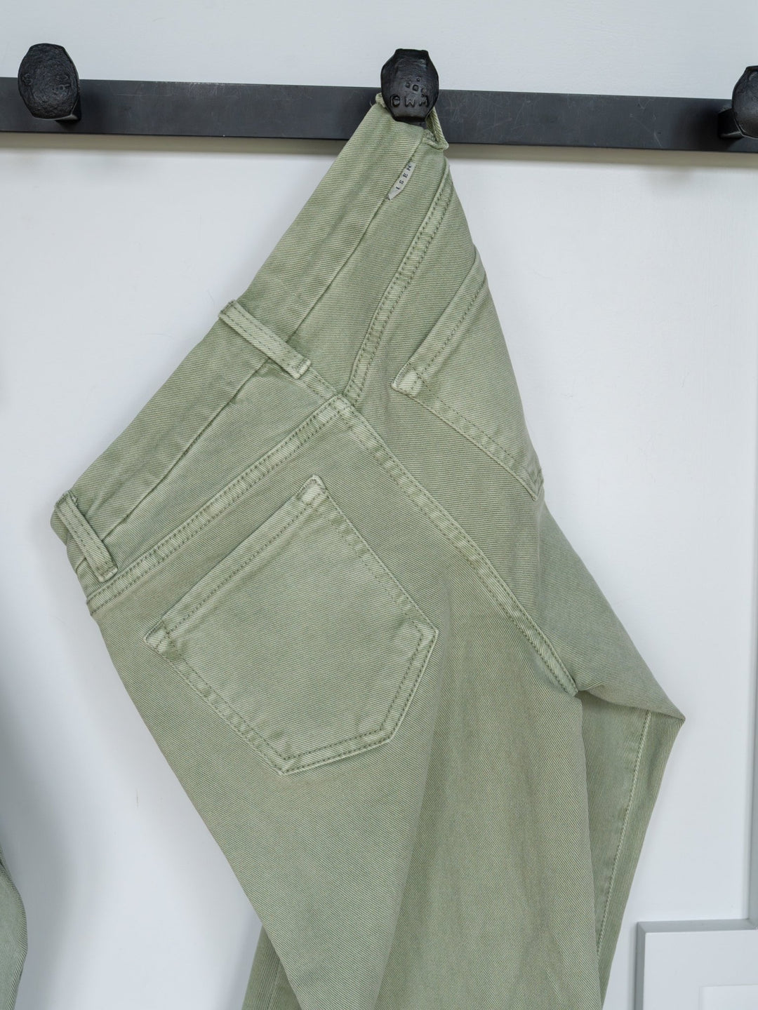 The Lindy High Rise Wide Leg Jean by Savannah Sevens western life{&}style hangs by a belt loop on a black hook against a white background, displaying the sage denim’s back pockets, seams, and raw cut hem.