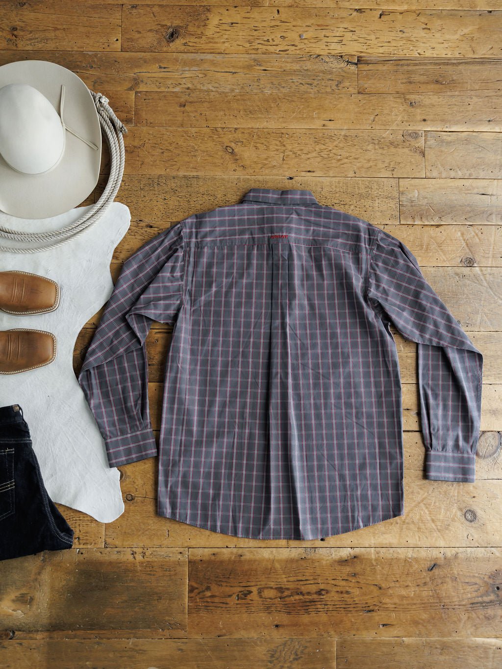 The Livingston Ariat Men's Grey Plaid Button Up by Savannah Sevens western life{&}style is styled laid flat with a white cowboy hat, tan boots, and dark jeans on a cowhide rug—a classic men’s shirt look.
