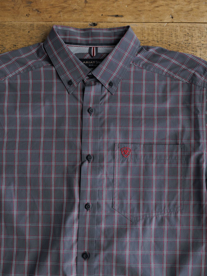 The Livingston Ariat Men's Grey Plaid Button Up by Savannah Sevens western life{&}style, featuring a button-down collar, chest pocket with red embroidered shield logo, is displayed flat on a wooden surface for classic plaid style.