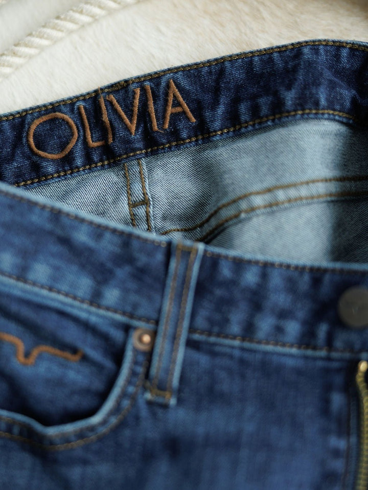 Close-up of The Livy High Rise 90's Wide Leg Jean *Exclusive* by Savannah Sevens western life{&}style, featuring premium blue denim with visible stitching, pocket detailing, and "OLIVIA" stitched in brown on the inside waistband.