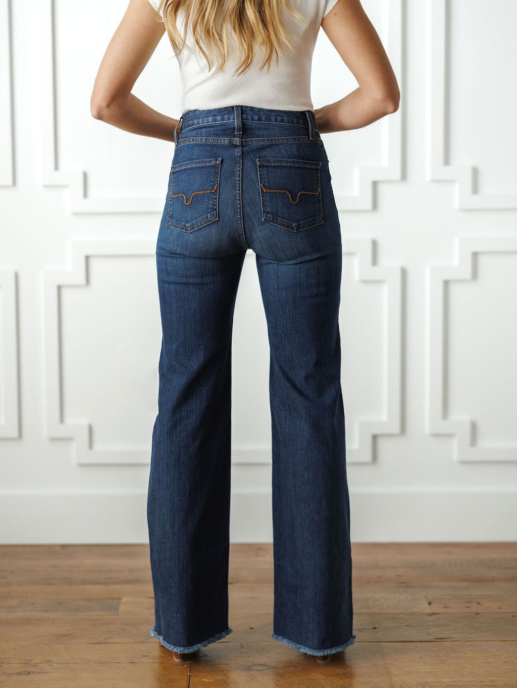 A woman stands indoors facing away, wearing a white short-sleeve top and Savannah Sevens western life{&}style The Livy High Rise 90’s Wide Leg Jean *Exclusive* with back pockets and frayed hems; her long blonde hair falls over her shoulders.