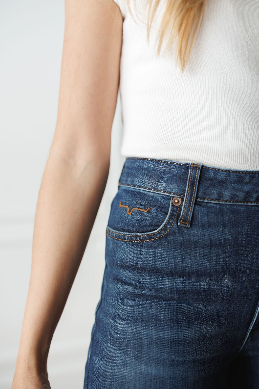 A person wears a white sleeveless top and Savannah Sevens western life{&}style’s The Livy High Rise 90's Wide Leg Jean *Exclusive*, featuring a straight cut, orange stitching, and a small embroidered pocket detail. Shown waist to mid-arm.