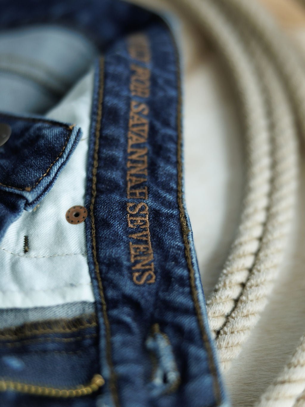 Close-up of The Livy High Rise 90's Wide Leg Jean *Exclusive* by Savannah Sevens western life{&}style, featuring a straight cut and brown "SAVANNAH SEVENS" stitching on the waistband, beside a coiled beige rope.