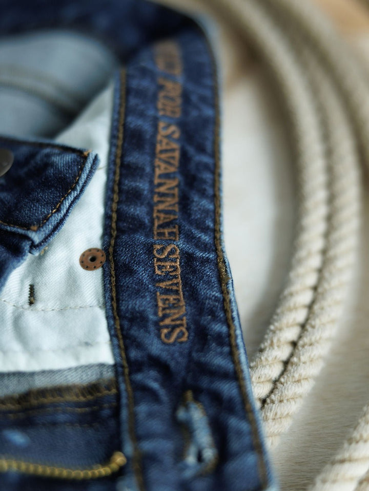Close-up of The Livy High Rise 90's Wide Leg Jean *Exclusive* by Savannah Sevens western life{&}style, featuring a straight cut and brown "SAVANNAH SEVENS" stitching on the waistband, beside a coiled beige rope.