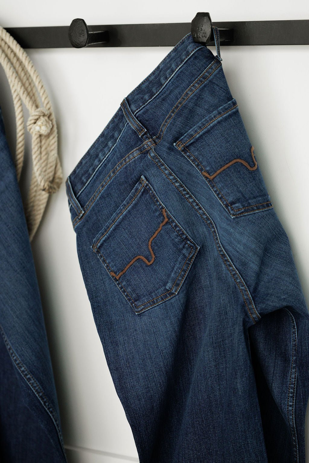 The Livy High Rise 90's Wide Leg Jean by Savannah Sevens western life{&}style hangs by the belt loop on a black wall hook, showcasing back pocket stitching. A rope is visible to the left.