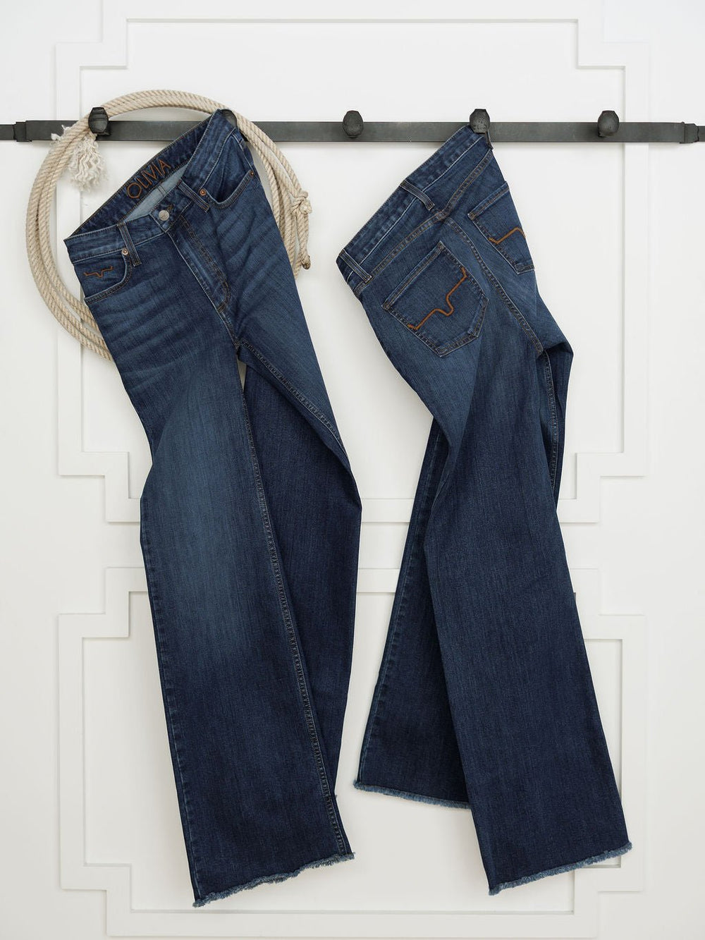 Two pairs of Savannah Sevens western life{&}style's The Livy High Rise 90's Wide Leg Jean *Exclusive* hang side by side on a black rod, with a coiled rope draped left, highlighting premium denim and classic Western style.