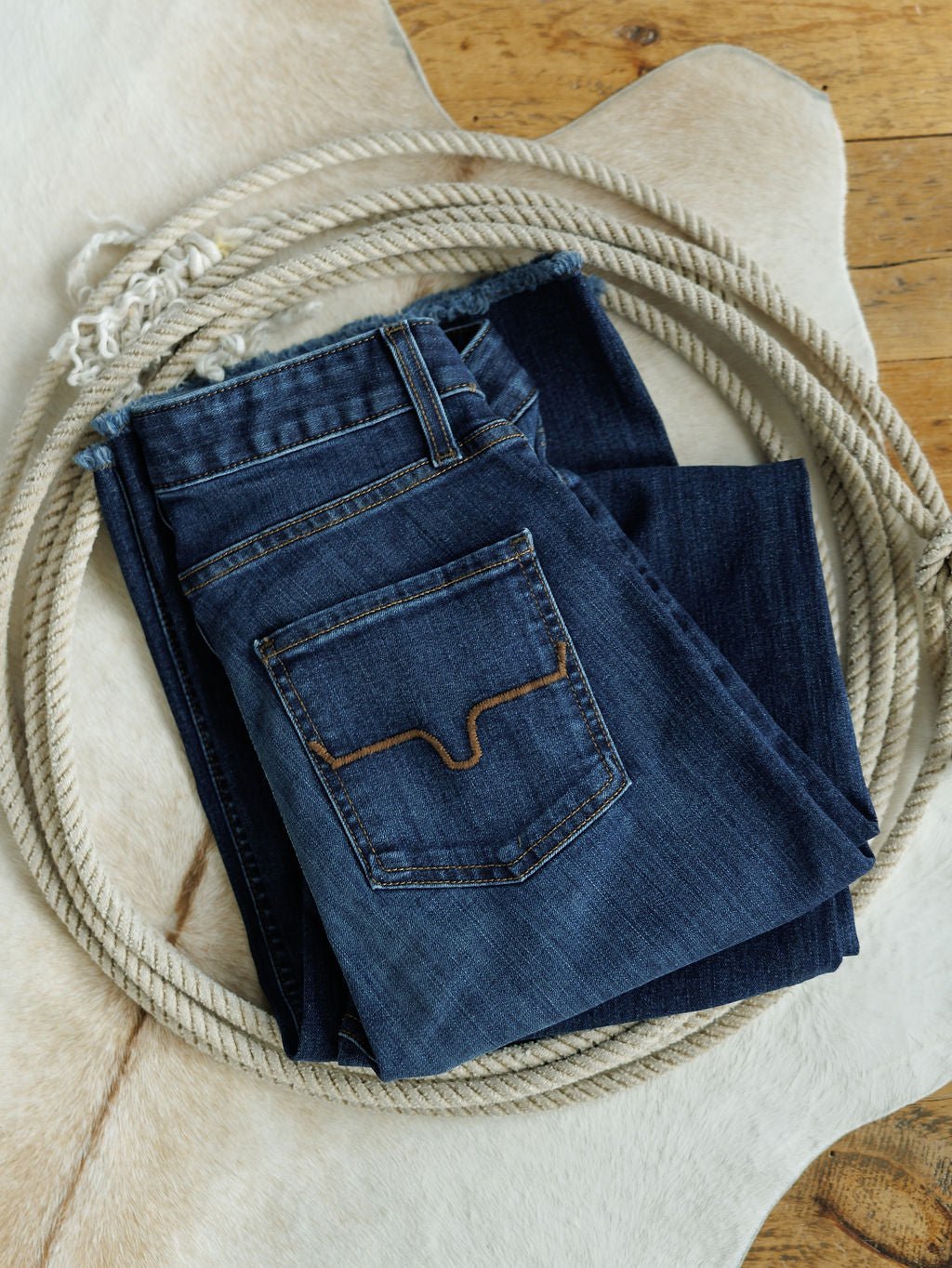 The Livy High Rise 90's Wide Leg Jean by Savannah Sevens western life{&}style, featuring brown-stitched back pockets, is displayed on a cowhide rug placed over a wooden floor.