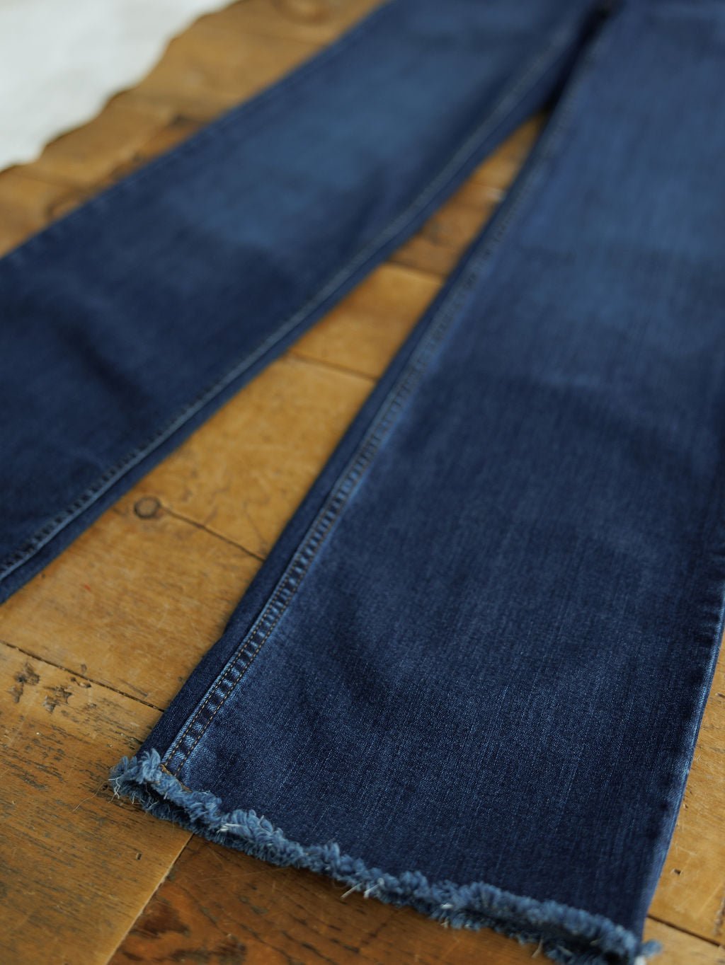 A close-up of The Livy High Rise 90's Wide Leg Jean *Exclusive* by Savannah Sevens western life{&}style, in dark blue premium denim with frayed hems, laid flat on a rustic wooden surface.