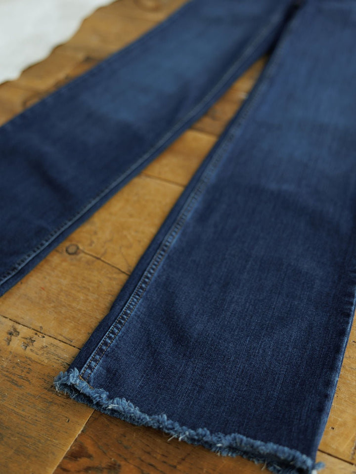 A close-up of The Livy High Rise 90's Wide Leg Jean *Exclusive* by Savannah Sevens western life{&}style, in dark blue premium denim with frayed hems, laid flat on a rustic wooden surface.