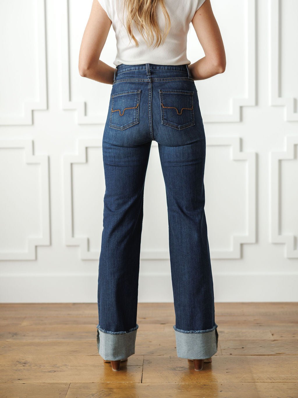 A woman stands indoors on a wooden floor by a white paneled wall, wearing Savannah Sevens western life{&}style's The Livy High Rise 90's Wide Leg Jean *Exclusive* and a white short-sleeve top.