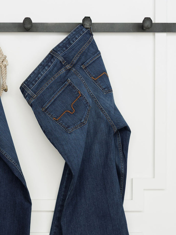 The Livy High Rise 90's Wide Leg Jean *Exclusive* by Savannah Sevens western life{&}style, in dark blue premium denim with brown stitching, hangs on a black wall hook against a white paneled wall, shown from the back to display the rear pockets and waistband.