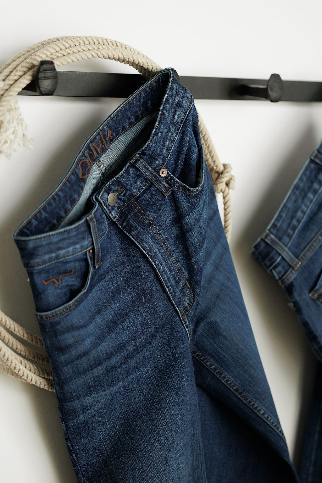 The Livy High Rise 90's Wide Leg Jean *Exclusive* by Savannah Sevens western life{&}style hangs on a black hook with coiled rope, next to another partially visible pair against a white background.