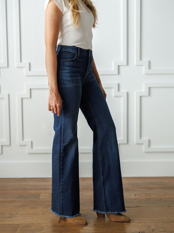 A woman stands indoors on a wooden floor against a white paneled wall, wearing a white sleeveless top and The Livy High Rise 90's Wide Leg Jean *Exclusive* by Savannah Sevens western life{&}style. Her face is not visible.