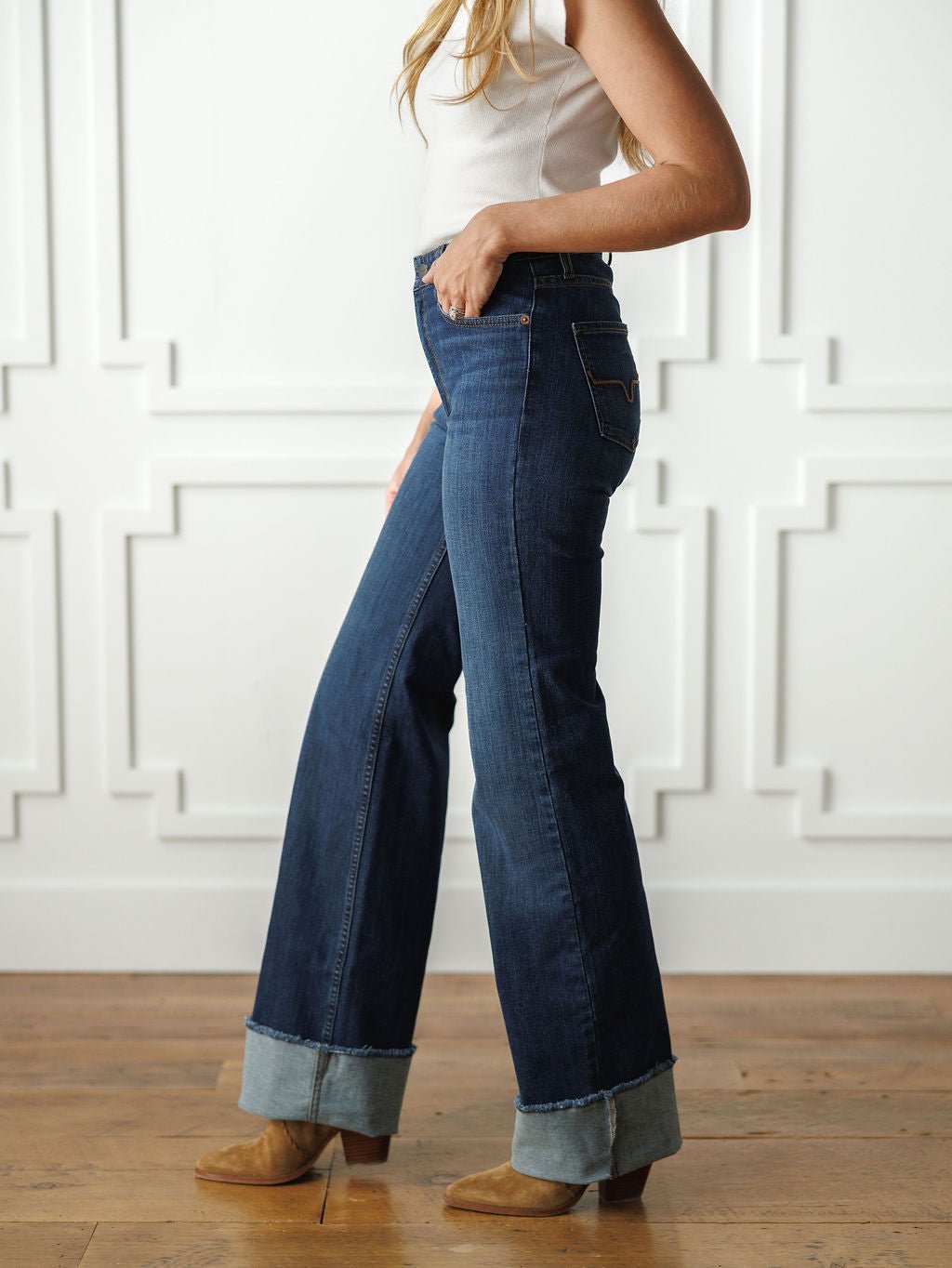 A woman in a white sleeveless top, Savannah Sevens western life{&}style’s The Livy High Rise 90's Wide Leg Jean *Exclusive* with large cuffs, and tan suede shoes stands sideways before a white paneled wall on a wooden floor.