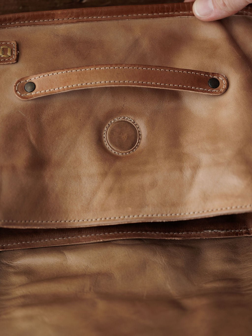 A close-up of the inside flap of the Savannah Sevens western life{&}style Lomita Leather Crossbody Bag showcases stitched circular detailing, two rivets, and a hand holding the edge to highlight its adjustable strap.