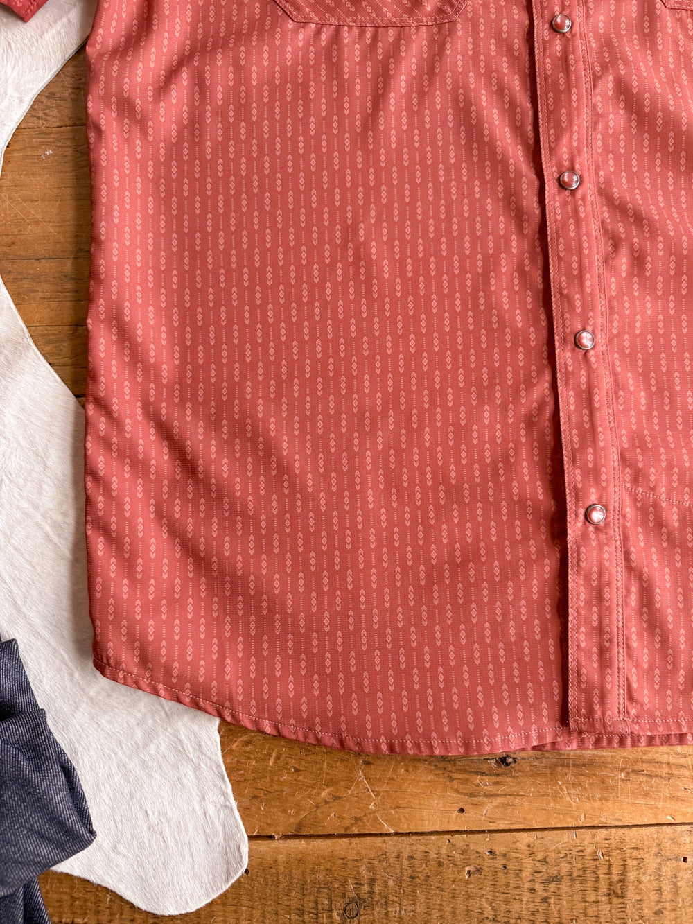 A close-up of the Longford Ariat Men’s Southwest Pearl Snap shirt by Savannah Sevens western life{&}style, featuring a salmon-pink pattern and buttons, lying on a wooden surface next to a white cowhide and dark blue fabric.