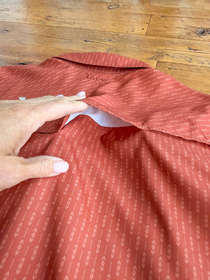 A hand lifts the collar of a patterned reddish-orange Longford Ariat Men’s Southwest Pearl Snap shirt from Savannah Sevens western life{&}style, revealing a hidden zipper pocket on the inside. The men’s pearl snap shirt is laid flat on a wooden floor.
