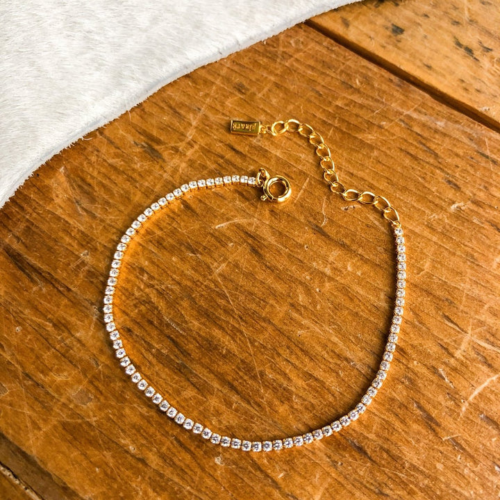 The Lorena Bracelet by Savannah Sevens western life{&}style, featuring small clear gemstones and an adjustable chain clasp, is shown in 18K gold plating on a wooden surface beside white fur fabric.