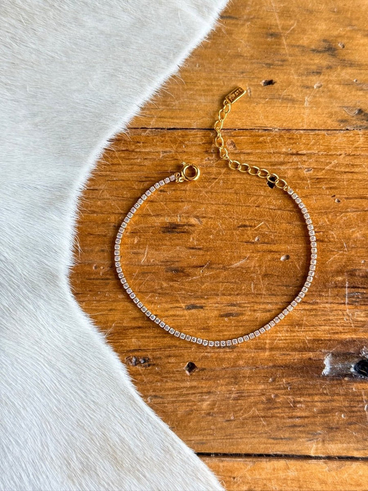 The Lorena Bracelet by Savannah Sevens western life{&}style, a delicate gold tennis bracelet with small clear cubic zirconia stones, is displayed in a circle on a wooden surface beside white fur or fabric.