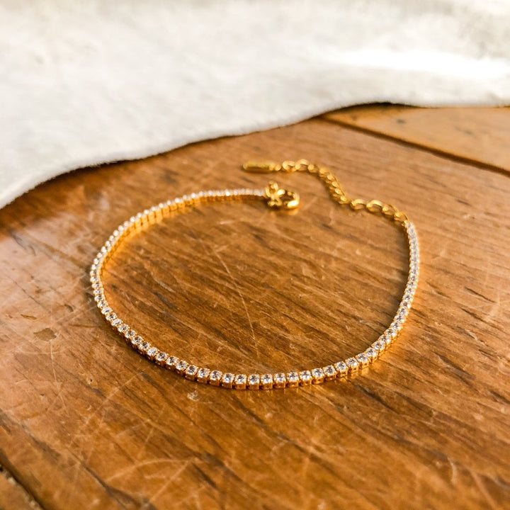 The Lorena Bracelet by Savannah Sevens western life{&}style, an 18K gold plated piece adorned with sparkling clear stones, is shown on a wooden surface with soft white fabric in the background.