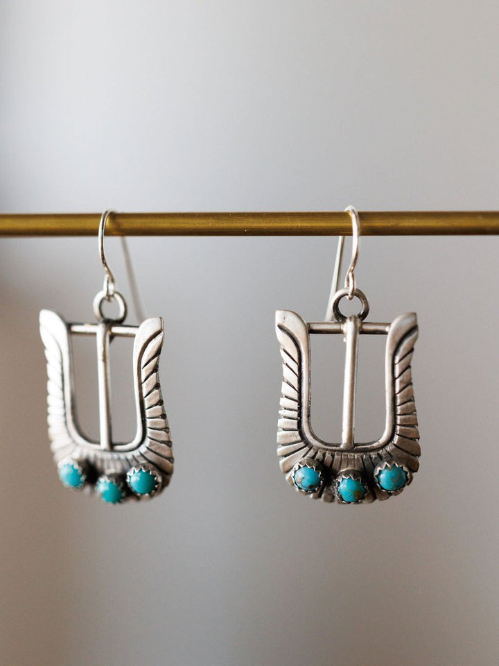 The Los Alamos Earrings by Savannah Sevens western life{&}style feature wing-inspired sterling silver with three turquoise stones, suspended from a gold-colored rod against a neutral background.