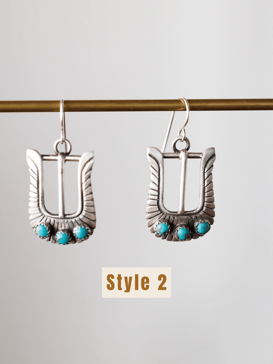 The Los Alamos Earrings by Savannah Sevens western life{&}style are sterling silver buckle-shaped earrings with three turquoise stones at the base, suspended from a horizontal rod. “Style 2” is shown below these unique turquoise earrings.