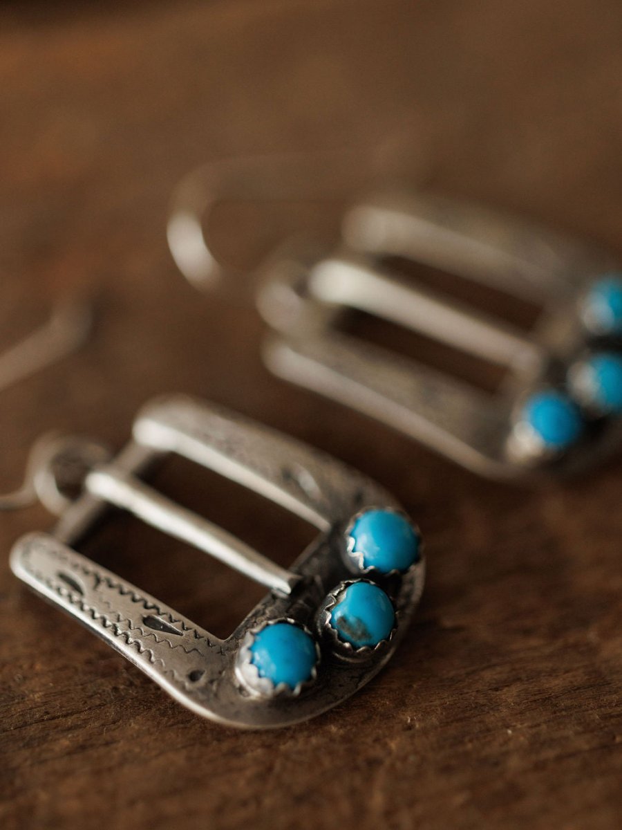Close-up of the Los Alamos Earrings by Savannah Sevens western life{&}style, sterling silver and shaped like horseshoe buckles, each set with two round turquoise stones at the bottom and displayed on a wooden surface. Another earring is softly blurred in the background.
