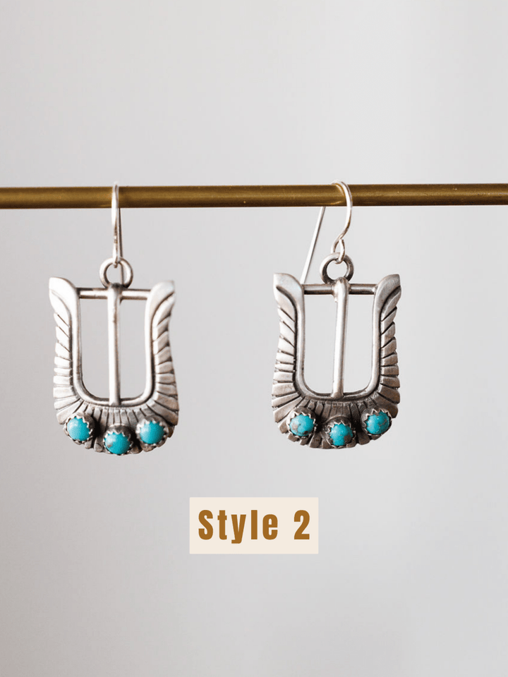 The Los Alamos Earrings by Savannah Sevens western life{&}style are sterling silver buckle-shaped earrings with three turquoise stones at the base, suspended from a horizontal rod. “Style 2” is shown below these unique turquoise earrings.