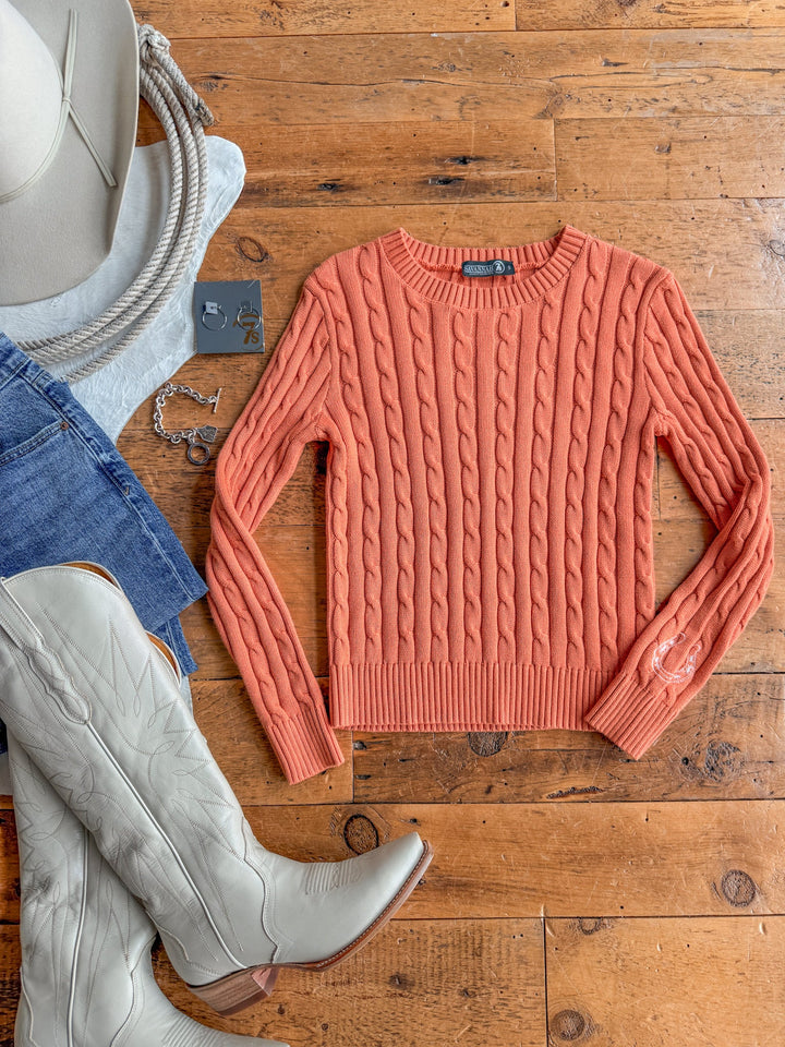 A Luck Orange Cable Knit Sweater {Exclusive} by Savannah Sevens western life{&}style, crafted from 100% cotton, is laid on a wooden floor and styled with gray cowboy boots, denim shorts, a rope, a gray hat, earrings, and a bracelet arranged around it.