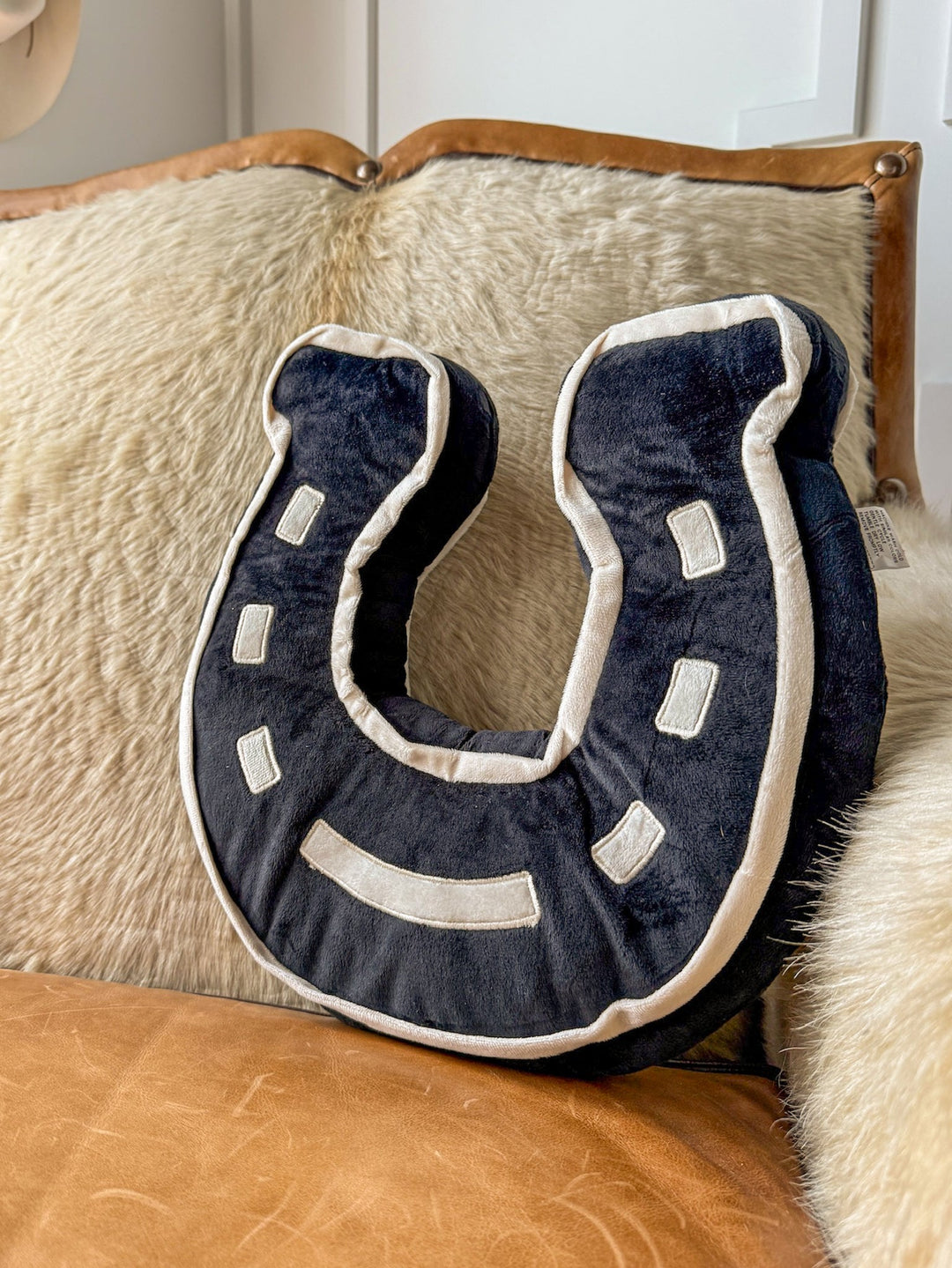 The Lucky Black Horseshoe Pillow by Savannah Sevens western life{&}style, showcasing black fabric with white detailing, rests on a tan leather couch against cream-colored cushions—an ideal accent for any western bedroom decor.
