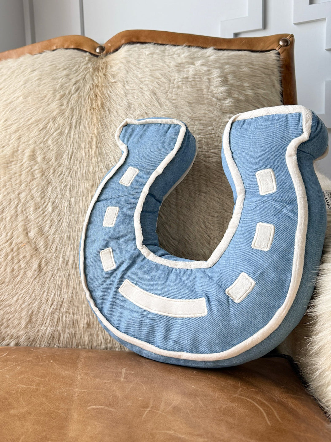 The Lucky Denim Horseshoe Pillow by Savannah Sevens western life{&}style, featuring denim blue and white accents, rests atop a tan fur cushion and leather seat—making it the perfect western bedroom accessory.
