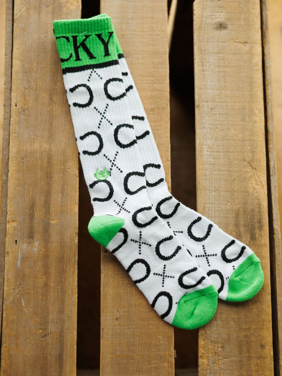 The Lucky Horseshoe Crew Socks by Savannah Sevens western life{&}style are white crew socks with green toes, heels, and cuffs. "LUCKY" is printed in green at the top, with black horseshoe and X patterns throughout.