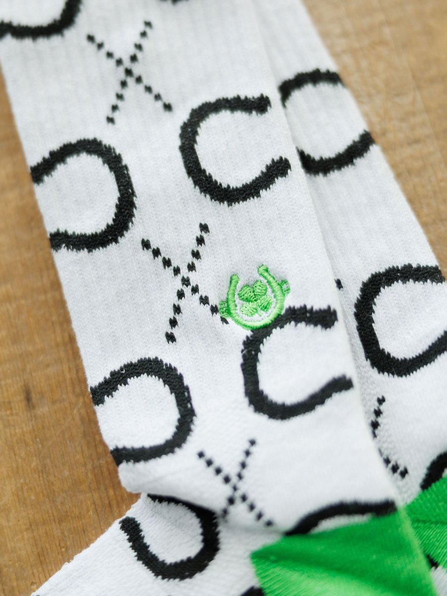 The Lucky Horseshoe Crew Socks by Savannah Sevens western life{&}style feature a white breathable design with black X and O patterns and a green embroidered snail on one sock, displayed on a wooden surface.