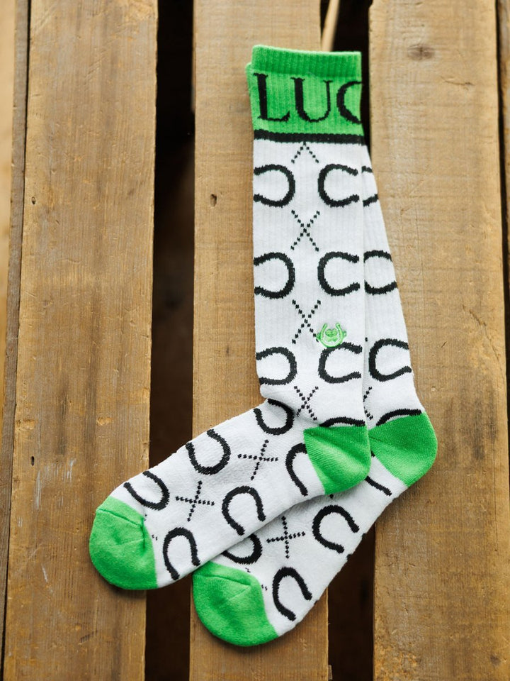 The Lucky Horseshoe Crew Socks by Savannah Sevens western life{&}style feature breathable fabric, green accents, black horseshoe and X designs, and "LUCK" on the cuff. They are displayed on a wooden surface.