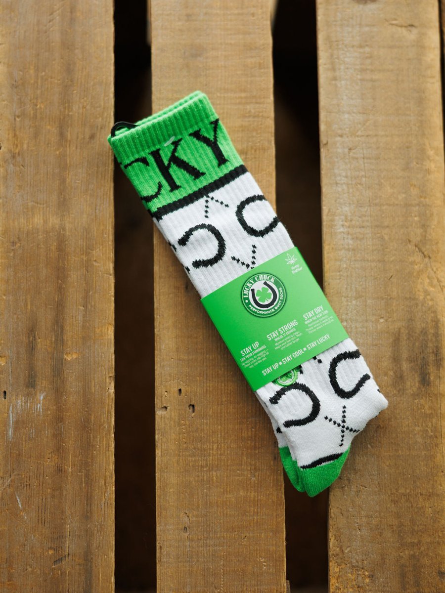 A pair of green and white Lucky Horseshoe Crew Socks by Savannah Sevens western life{&}style featuring a horseshoe pattern and "LUCKY" text are wrapped in green packaging and displayed on a wooden surface.