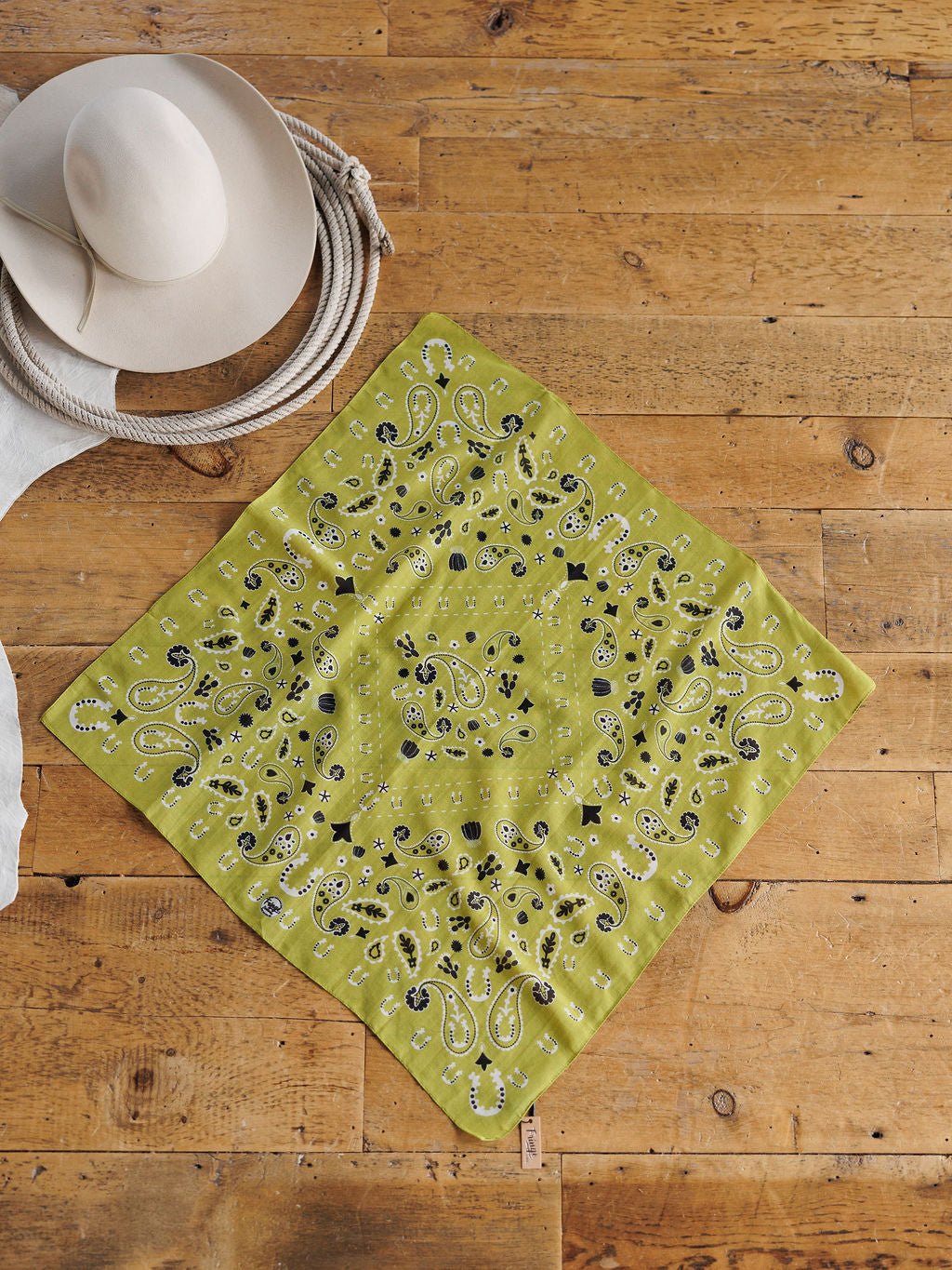 The Lucky Paisley Chartreuse Cotton-Silk Neck Scarf by Savannah Sevens western life{&}style is displayed on a wooden floor beside a coiled rope and a beige wide-brimmed hat.