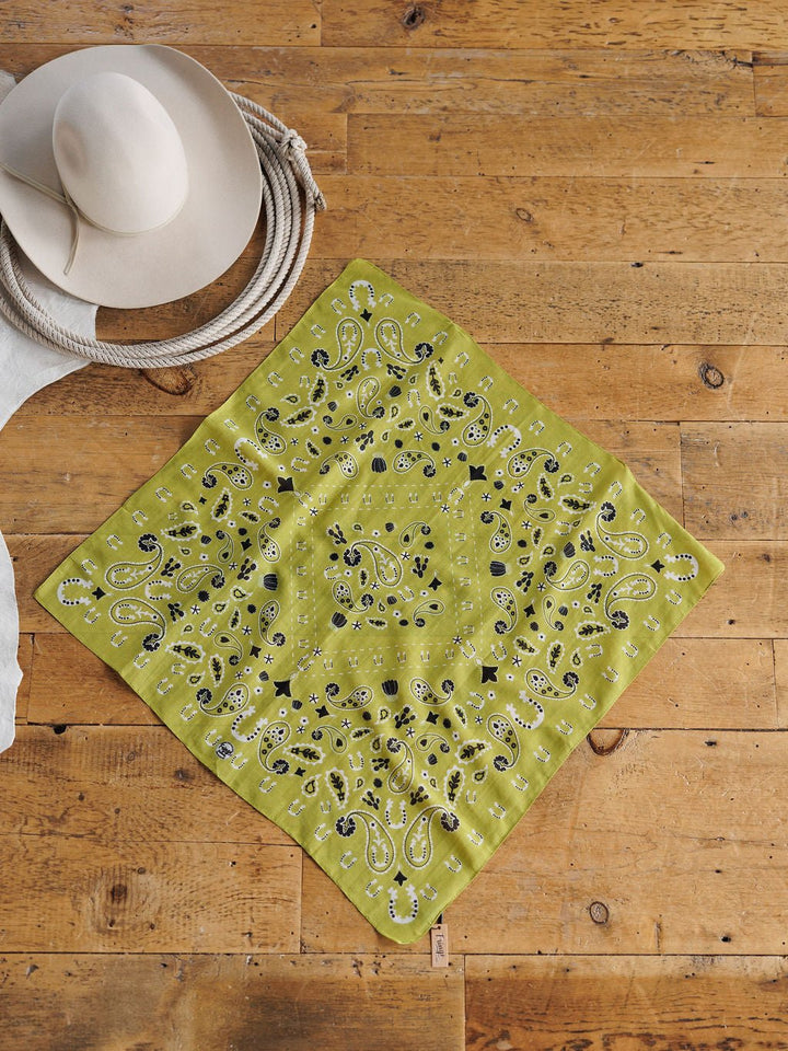 The Lucky Paisley Chartreuse Cotton-Silk Neck Scarf by Savannah Sevens western life{&}style is displayed on a wooden floor beside a coiled rope and a beige wide-brimmed hat.