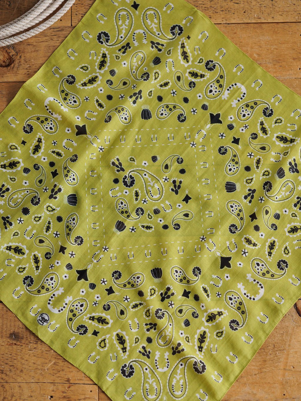 The Lucky Paisley Chartreuse Cotton-Silk Neck Scarf by Savannah Sevens western life{&}style, featuring a black and white paisley pattern, is spread out on a wooden surface with a coiled white rope in the top left corner.