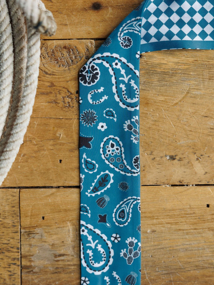 The Lucky Paisley Twilly {Cool Blue} by Savannah Sevens western life{&}style, featuring white and black paisley and floral patterns with a blue and white diamond-checkered underside, rests on wood beside a coiled rope.