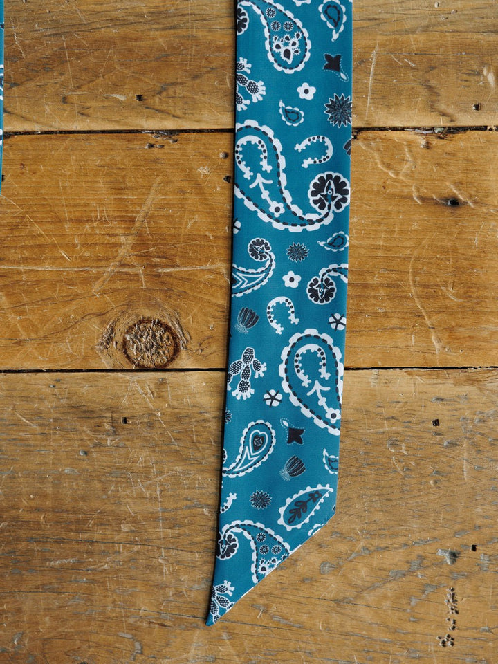 The Lucky Paisley Twilly {Cool Blue} by Savannah Sevens western life{&}style, featuring a teal paisley design with white and black accents, is displayed flat on a rustic wooden surface with visible grain and knots.