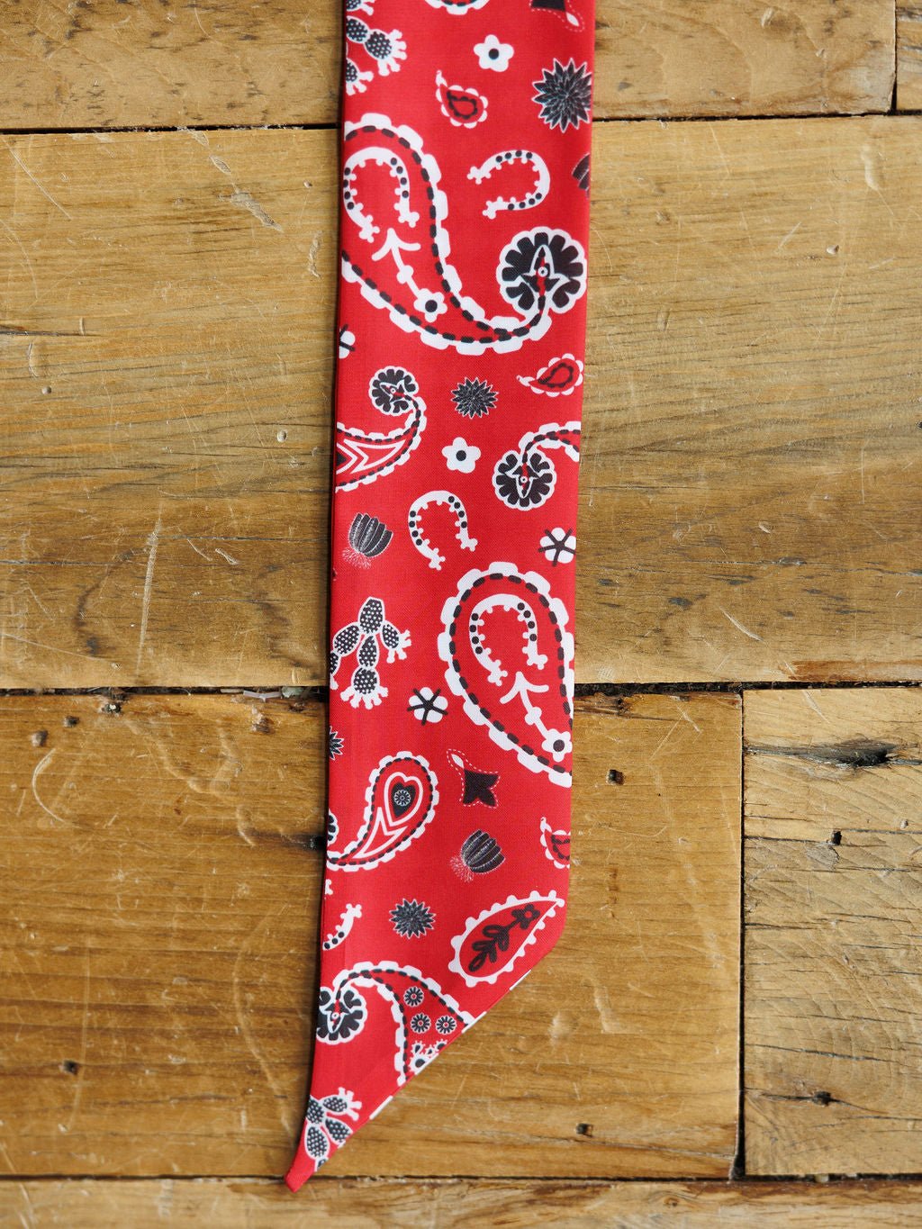The Lucky Paisley Twilly {Red} by Savannah Sevens western life{&}style, featuring a red satin scarf with black and white paisley graphics, rests on rustic light brown wooden planks.