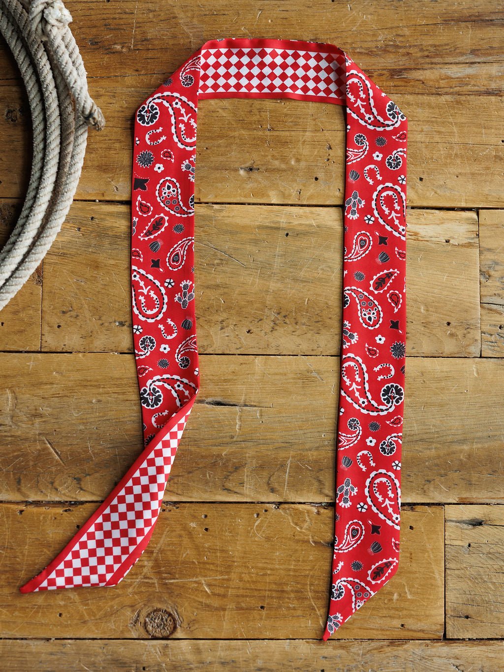The Lucky Paisley Twilly {Red} by Savannah Sevens western life{&}style features paisley on one side and red-and-white checks on the other, shown flat on wood beside a coiled rope.
