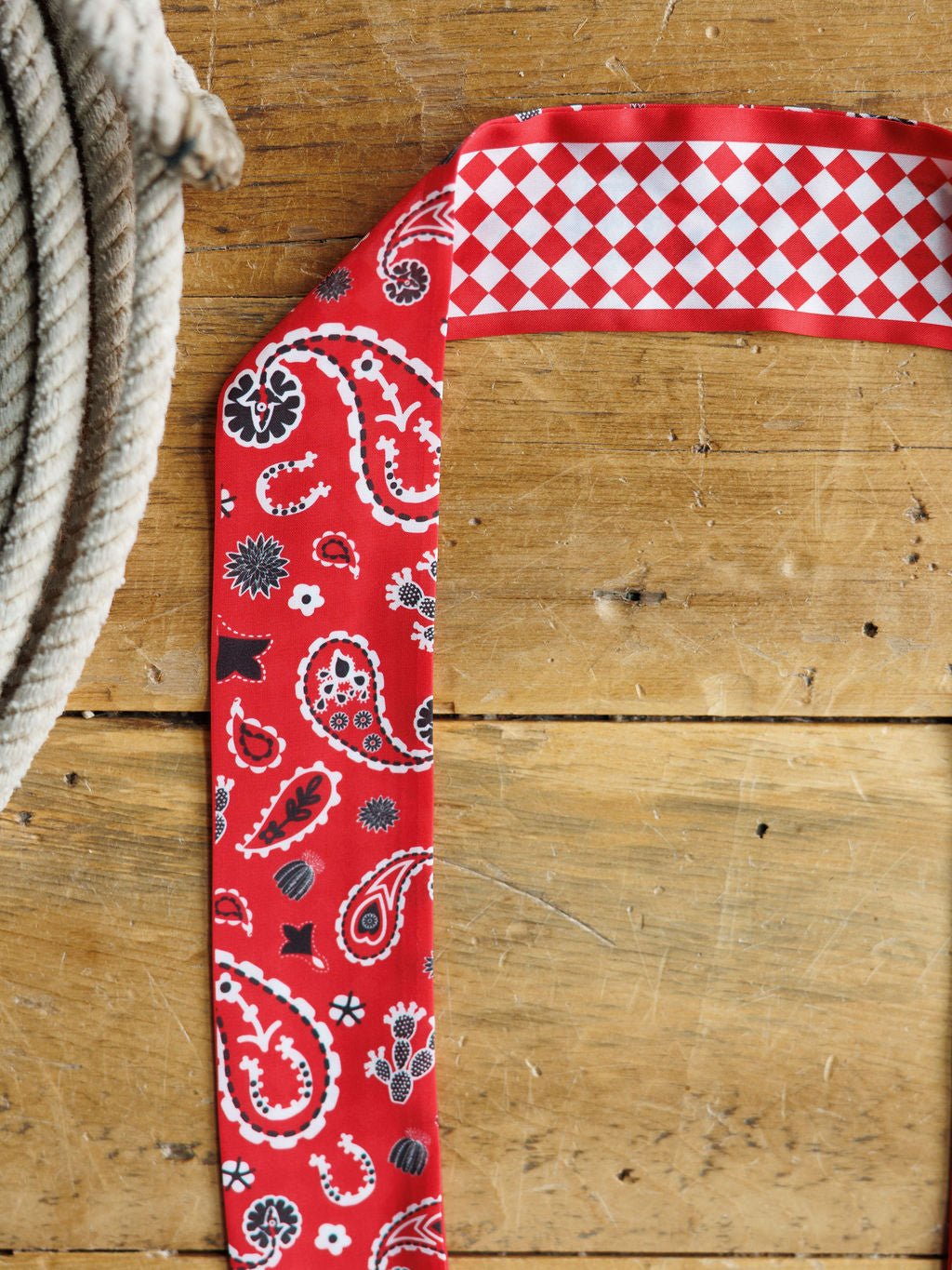 A coiled rope rests on wood next to the Savannah Sevens western life{&}style Lucky Paisley Twilly {Red}, which features a paisley pattern on one side and a red-and-white checkered design on the other.