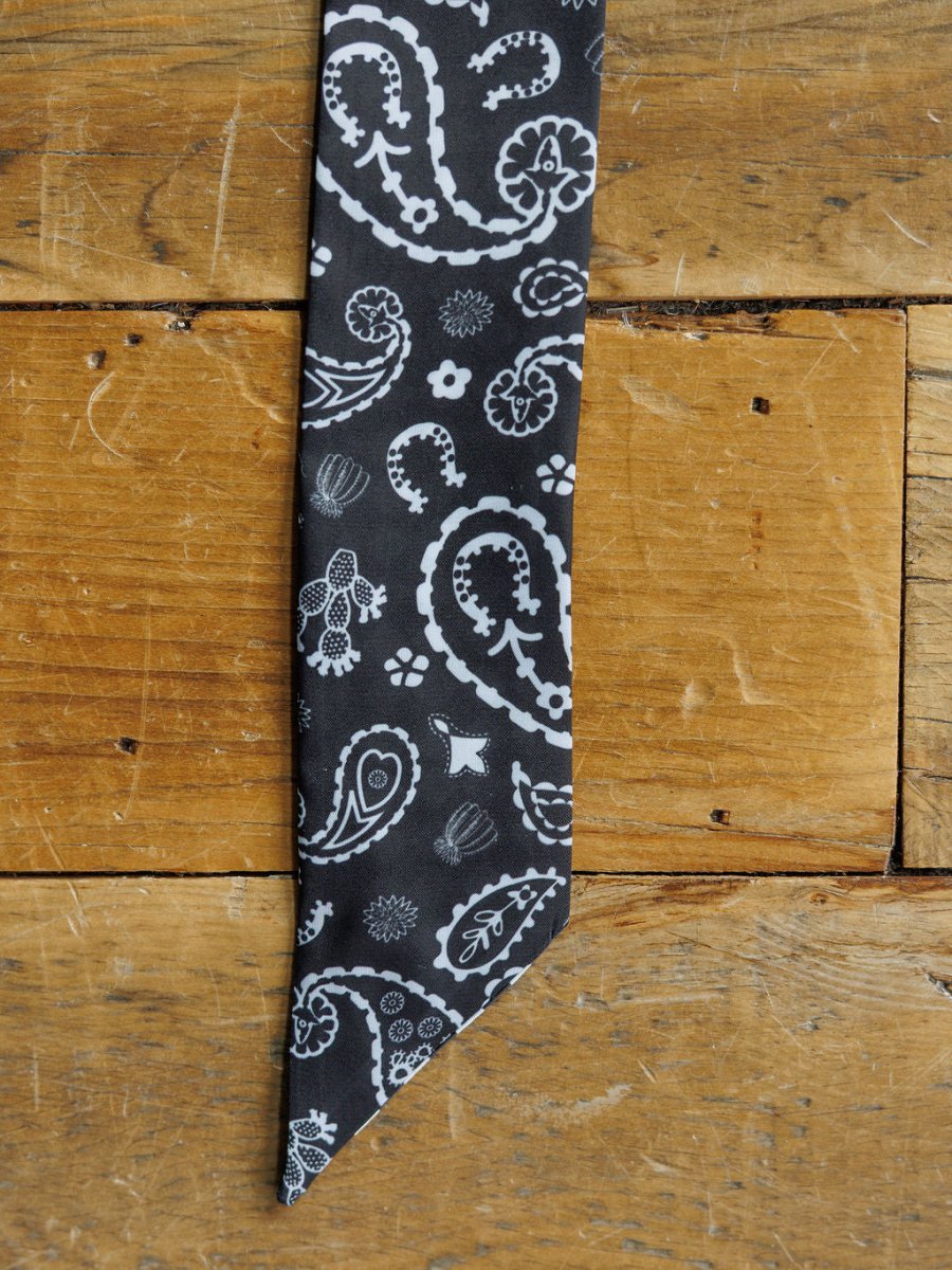 A close-up of the pointed end of the Lucky Paisley Twilly {Black} by Savannah Sevens western life{&}style, featuring a black satin fabric with white paisley and floral design, laid on a rustic wooden surface.