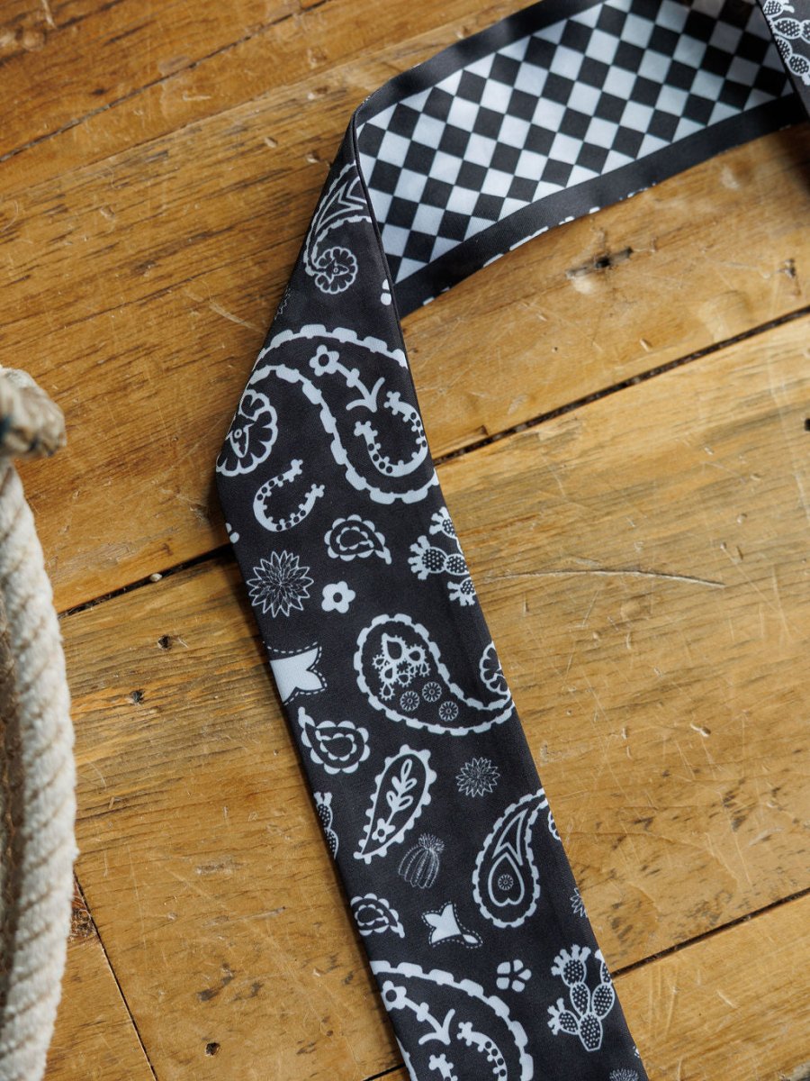 The Lucky Paisley Twilly {Black} by Savannah Sevens western life{&}style, featuring white paisley and floral patterns on black satin, is displayed on wood with its narrow end flipped to show a checker lining; a coiled rope appears at the left.