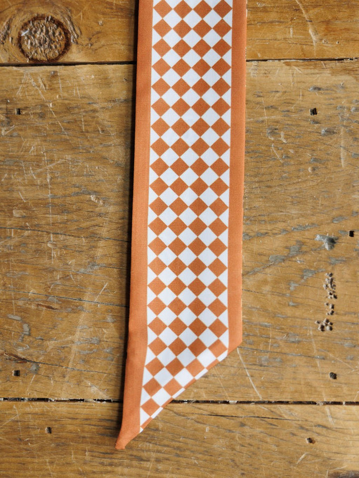 The Lucky Paisley Twilly {Rust} by Savannah Sevens western life{&}style, featuring a white and orange checkerboard pattern with an orange border, lies on a rustic wooden surface.